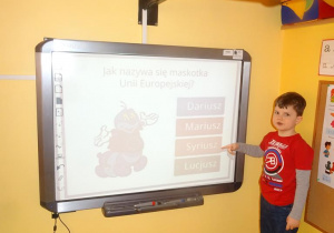 Chłopiec stoi pod tablicą interaktywną i wskazuje napis z nazwę maskotki Unii Europejskiej.